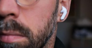 Photo en gros plan d'un homme barbu portant des écouteurs Apple AirPods blancs dans l'oreille gauche, illustrant les rumeurs des futurs AirPods Ultra prévus pour septembre 2026 avec caméra infrarouge intégrée et chip H3