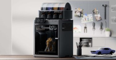Imprimante 3D Bambu Lab X2D posée sur un bureau en bois dans un atelier lumineux, avec son système AMS 2 multi-filaments en haut et une impression de sculpture en cours de réalisation dans la chambre