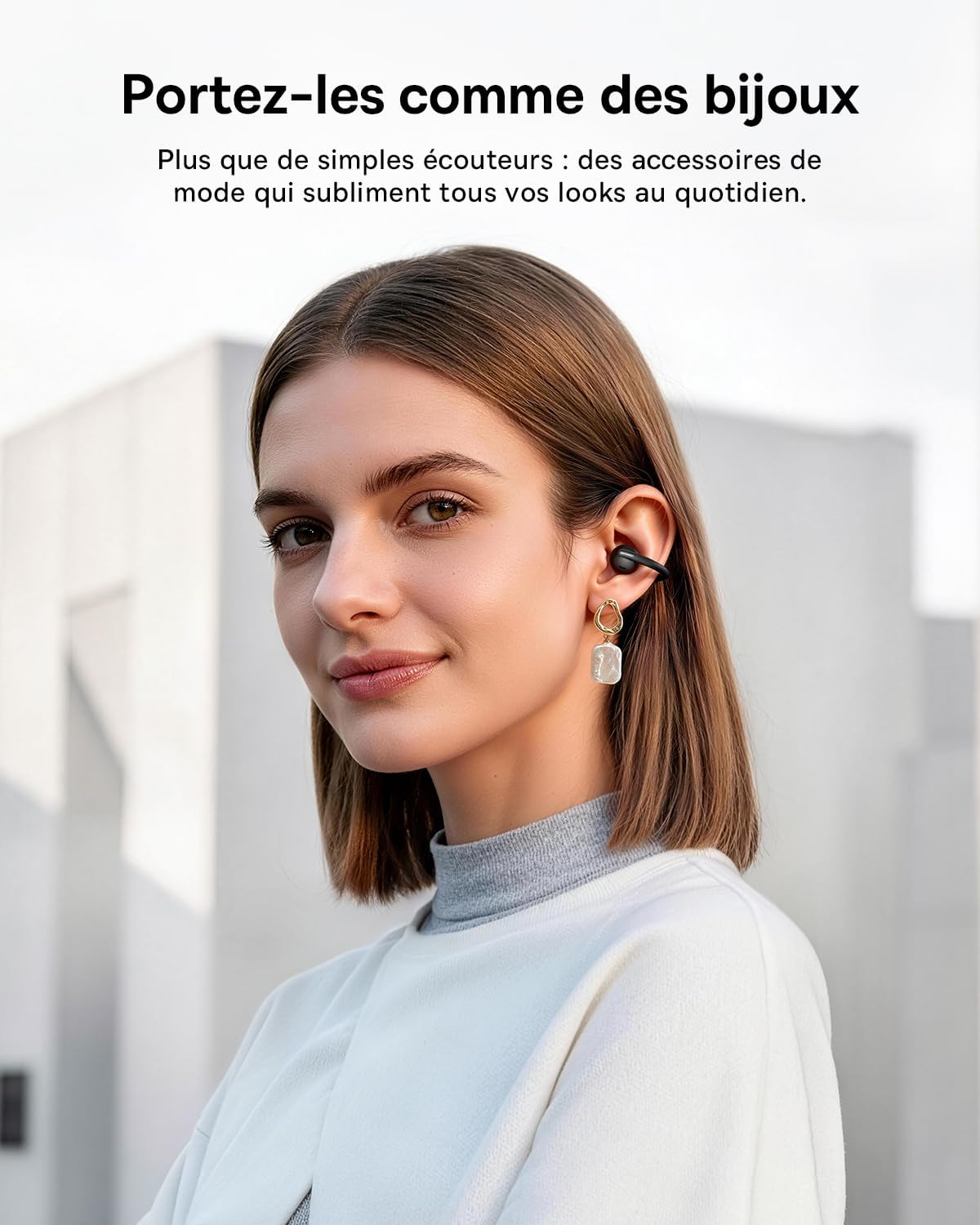 Jeune femme aux cheveux bruns courts portant un écouteur clip-on Baseus MC2 noir sur l'oreille droite, harmonieusement associé à une boucle d'oreille pendante en or et nacre, vêtue d'un haut blanc et col roulé gris clair, photographiée en portrait extérieur sur fond d'architecture moderne minimaliste avec slogan en français Portez-les comme des bijoux