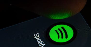 Gros plan sur un doigt s'apprêtant à cliquer sur l'icône de l'application Spotify sur un écran de smartphone rétroéclairé.