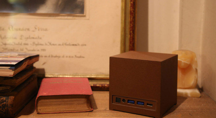Mini PC Kubb Essential de Bleujour en version chocolat/marron texturé, posé sur une étagère en bois rustique à côté de livres anciens. On distingue nettement la connectique frontale avec ses ports USB bleus. Le design cubique moderne contraste avec un cadre doré et des objets de décoration classiques en arrière-plan.