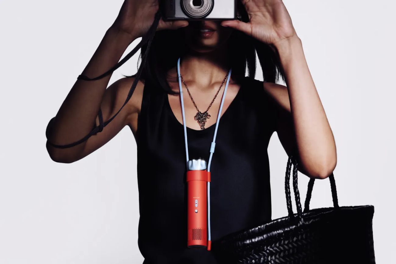 Gros plan d'une personne portant le ventilateur Dyson HushJet Mini Cool orange autour du cou comme un collier grâce à un cordon bleu clair. La personne tient un appareil photo argenté devant son visage et un sac tissé noir à la main, illustrant la portabilité et le style de l'appareil.