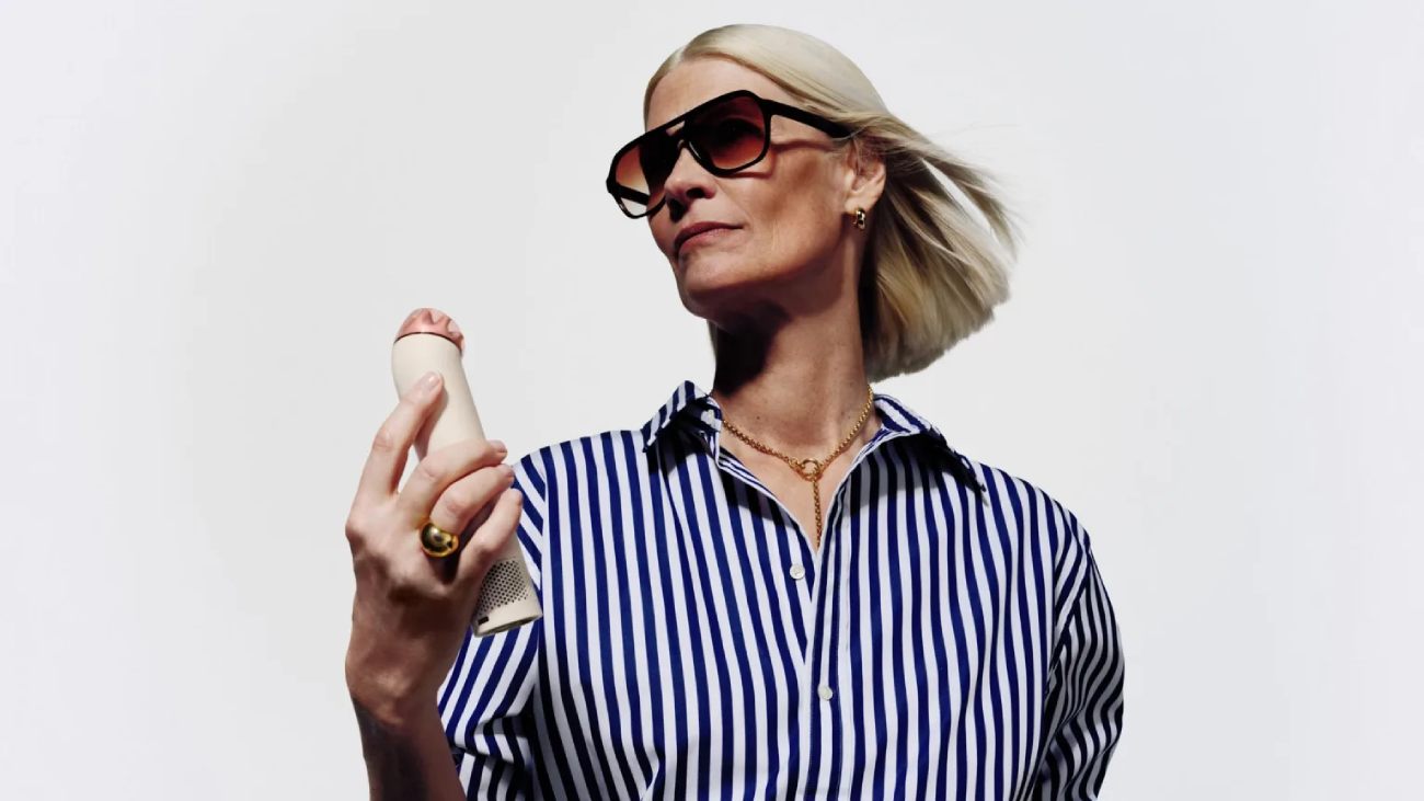 Femme blonde portant des lunettes de soleil et une chemise à rayures bleues, tenant le ventilateur portable Dyson HushJet Mini Cool en finition Stone Blush. Le produit compact est orienté vers son visage pour illustrer la technologie de flux d'air dirigé.
