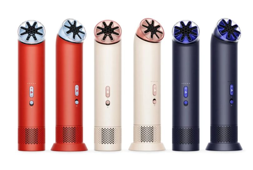Une femme élégante avec des lunettes de soleil aviateur regarde vers le haut tout en tenant le ventilateur portable Dyson HushJet Mini Cool de couleur Stone Blush, mettant en valeur son design compact et la technologie miniaturisée Air Multiplier.