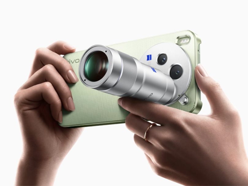 Gros plan sur un concept de Vivo X500 Pro Max vert pastel, tenu en main par un utilisateur avec une bague, arborant un module photo hybride unique : deux objectifs principaux avec logo ZEISS bleus et un objectif amovible/télescopique argenté proéminent.