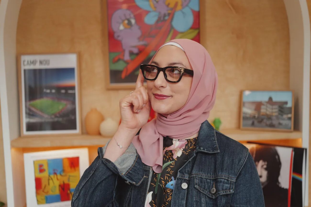Une femme portant un hijab rose et une veste en jean porte des lunettes intelligentes à monture noire, posant son index sur la branche pour suggérer l'interaction, dans un environnement de bureau moderne avec des affiches.