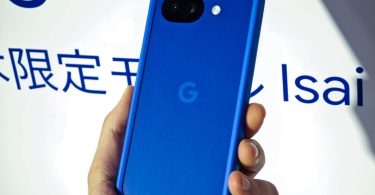 Gros plan sur le Google Pixel 10a Isai Blue en version japonaise. Le smartphone bleu électrique est tenu en main devant un panneau d'exposition affichant le texte "Isai". On distingue le double capteur photo arrière et le logo Google.