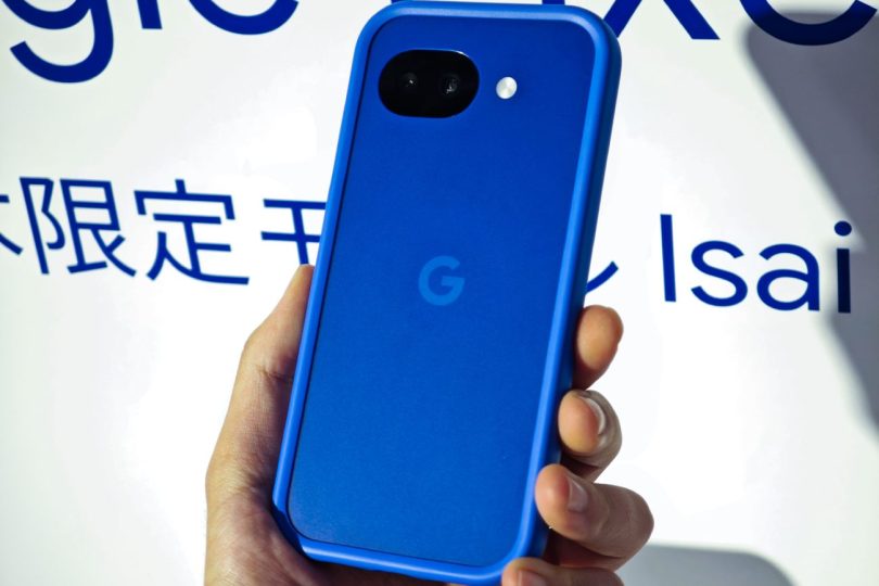 Gros plan sur le Google Pixel 10a Isai Blue en version japonaise. Le smartphone bleu électrique est tenu en main devant un panneau d'exposition affichant le texte "Isai". On distingue le double capteur photo arrière et le logo Google.