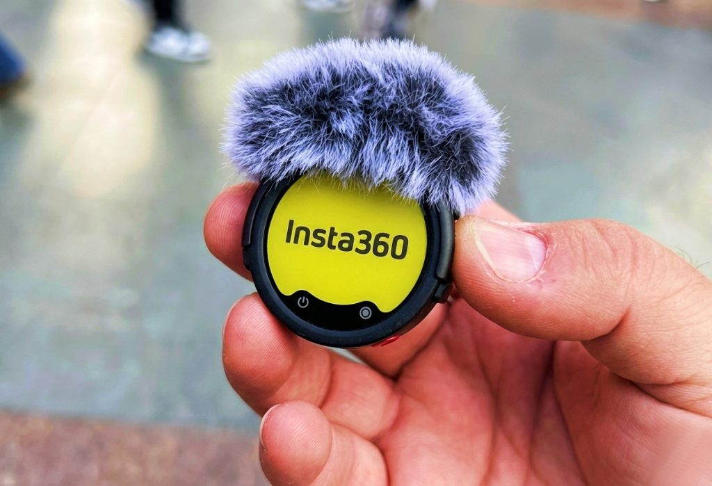 Vue rapprochée d'une main tenant l'émetteur circulaire de l'Insta360 Mic Pro, montrant l'écran E-Ink affichant "Insta360" sur fond jaune et une bonnette anti-vent grise.