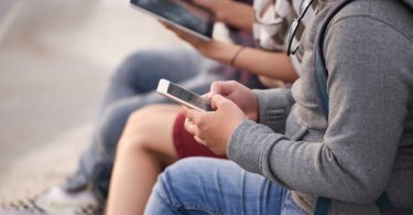 Trois adolescents assis à l'extérieur, concentrés sur leurs écrans. L'un au premier plan tape sur son smartphone, tandis qu'un autre, au second plan flou, tient une tablette. Ils portent des vêtements décontractés et des sacs à dos.