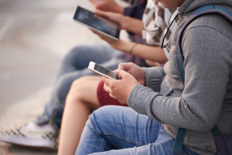 Trois adolescents assis à l'extérieur, concentrés sur leurs écrans. L'un au premier plan tape sur son smartphone, tandis qu'un autre, au second plan flou, tient une tablette. Ils portent des vêtements décontractés et des sacs à dos.