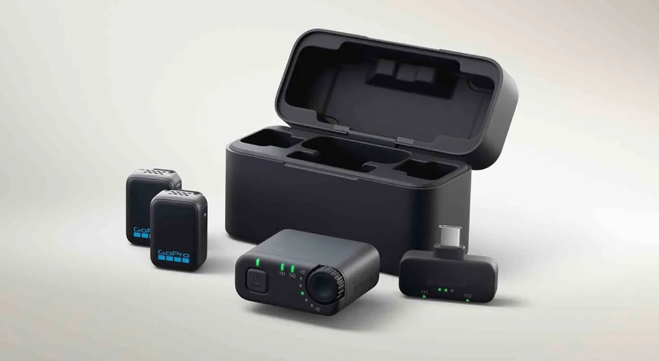 Ensemble complet du système de microphones sans fil GoPro pour la caméra Mission 1. On aperçoit deux émetteurs compacts avec logo GoPro, un boîtier de charge ouvert, un récepteur avec molette de gain et indicateurs LED verts, ainsi qu'un adaptateur USB-C. Design minimaliste noir sur fond clair.