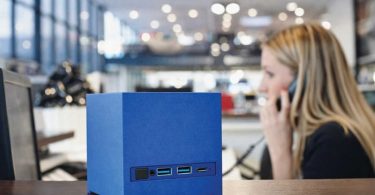Gros plan sur le mini PC français Kubb Essential de Bleujour en coloris bleu posé sur un comptoir. La face avant révèle le bouton d'alimentation et la connectique incluant deux ports USB-A et un port USB-C. En arrière-plan, une femme travaille dans un environnement de bureau lumineux.