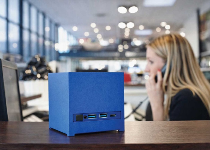 Gros plan sur le mini PC français Kubb Essential de Bleujour en coloris bleu posé sur un comptoir. La face avant révèle le bouton d'alimentation et la connectique incluant deux ports USB-A et un port USB-C. En arrière-plan, une femme travaille dans un environnement de bureau lumineux.
