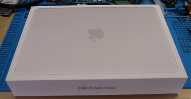 Boîte de vente blanche personnalisée avec l'inscription "MacBook Neo" et le logo Apple, posée sur un établi de réparation électronique bleu parsemé de composants et d'outils.