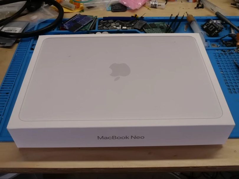 Boîte de vente blanche personnalisée avec l'inscription "MacBook Neo" et le logo Apple, posée sur un établi de réparation électronique bleu parsemé de composants et d'outils.