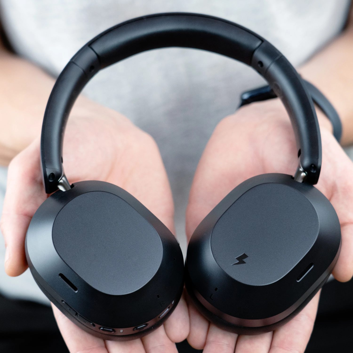 Vue de détail des deux coques du casque MEE Audio Matrix Cinema 3D tenu dans les mains, avec à gauche l'oreillette comportant le pictogramme éclair indiquant la zone de charge sans fil magnétique et un bouton physique dédié, et à droite l'oreillette comportant la surface tactile pour les gestes touch controls et trois boutons physiques alignés en bas pour le contrôle du volume et de la lecture