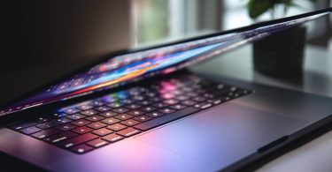 Gros plan sur un ordinateur portable noir dont l'écran, affichant des couleurs vives, est partiellement refermé, projetant des reflets colorés et des ombres sur le clavier et le trackpad. Un éclairage dramatique accentue la texture des touches dans un environnement de bureau sombre avec une plante en arrière-plan.