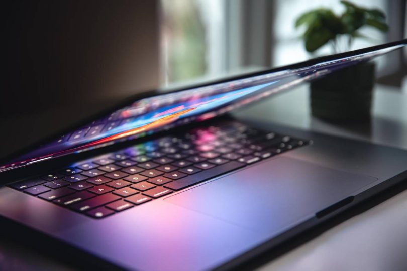 Gros plan sur un ordinateur portable noir dont l'écran, affichant des couleurs vives, est partiellement refermé, projetant des reflets colorés et des ombres sur le clavier et le trackpad. Un éclairage dramatique accentue la texture des touches dans un environnement de bureau sombre avec une plante en arrière-plan.