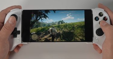 Vue rapprochée des mains d'un utilisateur tenant le smartphone RedMagic 11 Pro Golden Saga inséré dans une manette blanche de type télescopique. L'écran affiche une scène de Red Dead Redemption 2 en plein champ, avec un menu de statistiques de performance visible dans le coin supérieur gauche (GPU, CPU, RAM, FPS) et le personnage principal Arthur Morgan de dos.