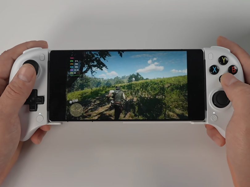 Vue rapprochée des mains d'un utilisateur tenant le smartphone RedMagic 11 Pro Golden Saga inséré dans une manette blanche de type télescopique. L'écran affiche une scène de Red Dead Redemption 2 en plein champ, avec un menu de statistiques de performance visible dans le coin supérieur gauche (GPU, CPU, RAM, FPS) et le personnage principal Arthur Morgan de dos.
