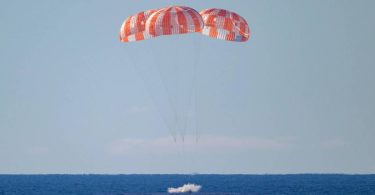 Une photographie de la capsule spatiale Orion de la NASA amerrissant dans l'Océan Pacifique. Elle est suspendue sous ses trois immenses parachutes principaux rayés d'orange et de blanc, qui sont entièrement déployés. La capsule vient de toucher l'eau, créant une petite gerbe d'écume blanche. La scène se déroule sous un ciel bleu clair, avec l'horizon de l'océan bleu s'étendant à l'arrière-plan.