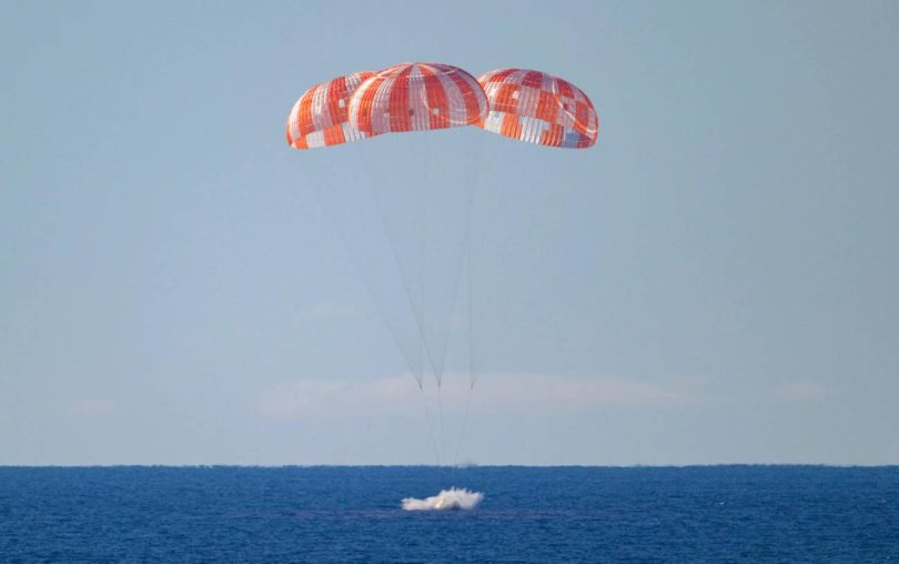 Une photographie de la capsule spatiale Orion de la NASA amerrissant dans l'Océan Pacifique. Elle est suspendue sous ses trois immenses parachutes principaux rayés d'orange et de blanc, qui sont entièrement déployés. La capsule vient de toucher l'eau, créant une petite gerbe d'écume blanche. La scène se déroule sous un ciel bleu clair, avec l'horizon de l'océan bleu s'étendant à l'arrière-plan.