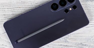 Description : Vue rapprochée de la face arrière d'un smartphone Samsung Galaxy S26 Ultra en coloris Titanium Violet, posé sur un support en bois blanc. Le module photo à cinq capteurs est visible en haut à droite, et le stylet S Pen gris est déposé en diagonale sur le châssis.