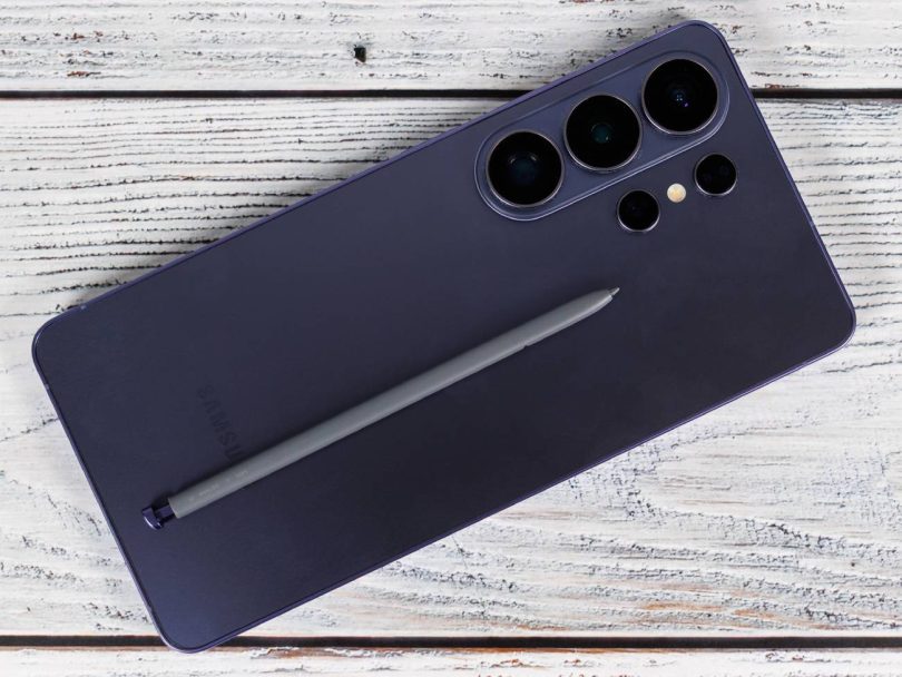 Description : Vue rapprochée de la face arrière d'un smartphone Samsung Galaxy S26 Ultra en coloris Titanium Violet, posé sur un support en bois blanc. Le module photo à cinq capteurs est visible en haut à droite, et le stylet S Pen gris est déposé en diagonale sur le châssis.