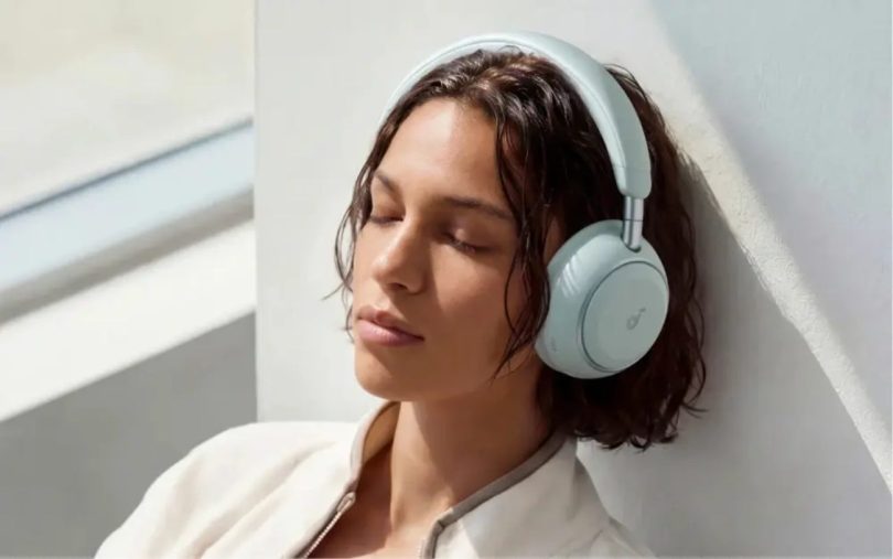 Femme aux cheveux bruns courts portant le casque Soundcore Space 2 en finition Seafoam Green vert pastel, yeux fermés, adossée à un mur clair éclairé par la lumière naturelle