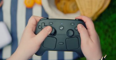 Prise de vue en plongée de mains tenant un Steam Controller noir en situation de jeu, sur un fond flou de pique-nique en extérieur avec nappe rayée et oranges.