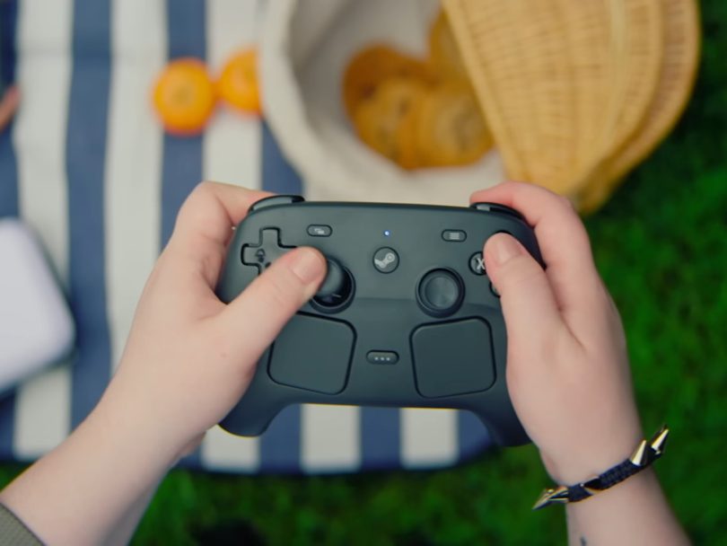 Prise de vue en plongée de mains tenant un Steam Controller noir en situation de jeu, sur un fond flou de pique-nique en extérieur avec nappe rayée et oranges.