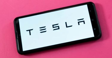 Photographie d'un smartphone noir posé sur un fond rose vif, affichant le logo noir de Tesla (le nom en toutes lettres) sur un écran blanc uni.