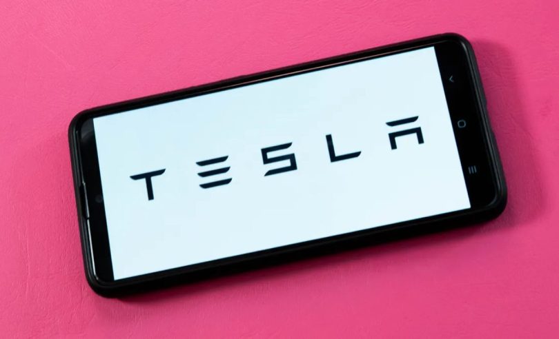 Photographie d'un smartphone noir posé sur un fond rose vif, affichant le logo noir de Tesla (le nom en toutes lettres) sur un écran blanc uni.