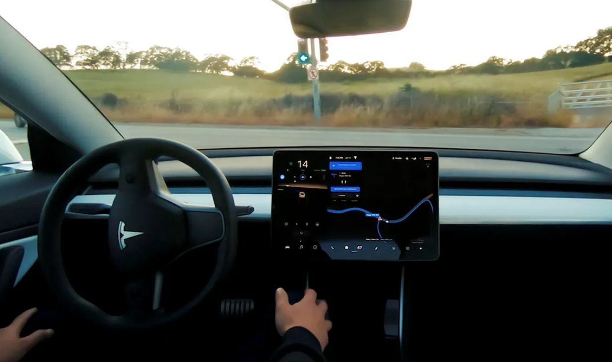 Vue de l'intérieur d'un véhicule Tesla (modèle Cybercab) montrant le volant, le grand écran central affichant une navigation cartographique et l'environnement extérieur de collines au crépuscule. La main du conducteur est visible.