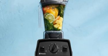 Description : Blender Vitamix Explorian E310 noir rempli de fruits frais (agrumes, mangue) et d'épinards sur un fond bleu clair, illustrant le modèle d'entrée de gamme dont la production s'arrête.