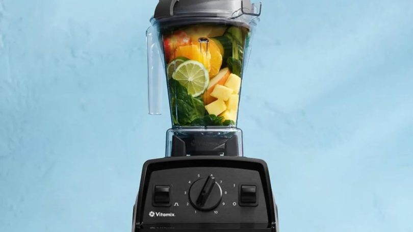 Description : Blender Vitamix Explorian E310 noir rempli de fruits frais (agrumes, mangue) et d'épinards sur un fond bleu clair, illustrant le modèle d'entrée de gamme dont la production s'arrête.