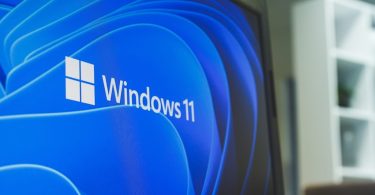 Description : Gros plan sur l'écran d'un ordinateur portable affichant le logo officiel et le texte "Windows 11" sur un fond bleu abstrait et moderne.