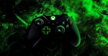 Manette Xbox noire avec logo et croix directionnelle rétroéclairés en vert néon, sur un fond de fumée émeraude stylisée évoquant l'identité visuelle historique de la marque.