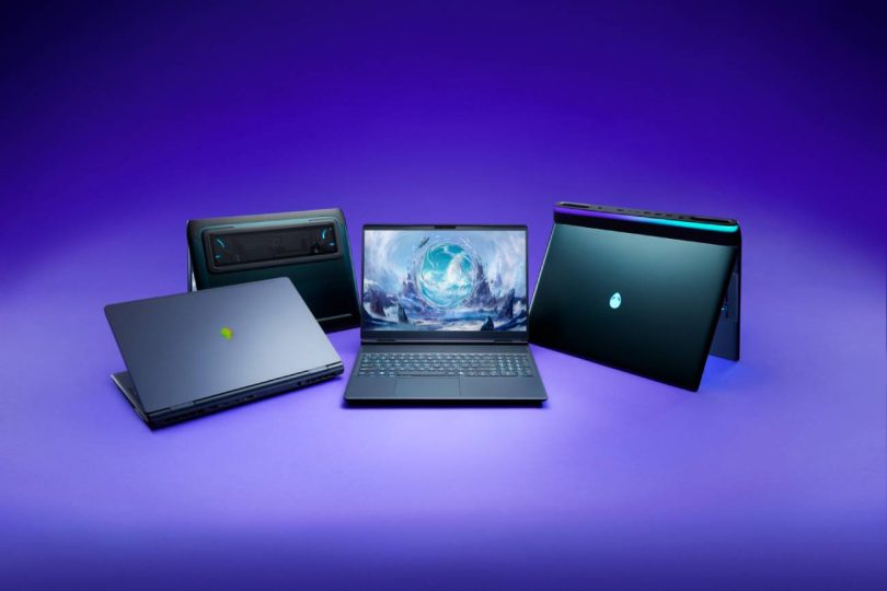 Quatre ordinateurs portables gaming Alienware disposés sur un fond violet épuré, mettant en valeur leurs lignes futuristes, le logo de la tête d'alien illuminé et un écran central affichant une illustration fantastique.