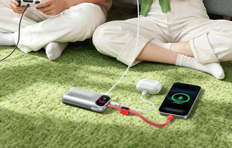 Batterie externe Ugreen PB610 45W argentée au design incurvé, dotée d'un écran affichant un emoji, posée sur un tapis vert et chargeant simultanément un iPhone et des écouteurs via son câble rouge tressé intégré.