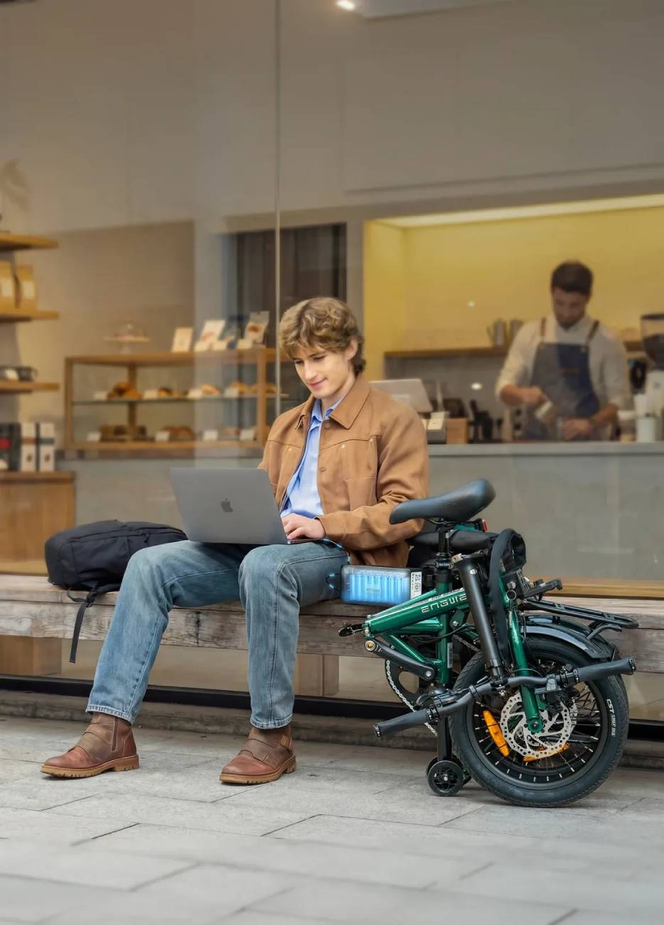 Un cycliste travaillant sur un ordinateur portable devant un café, avec son vélo électrique pliable Engwe Zip replié et la batterie amovible de 360Wh rétroéclairée en bleu posée sur le banc.