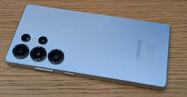 Dos d'un smartphone Samsung Galaxy S26 bleu clair avec ses capteurs photo texturés, posé sur une table en bois, illustrant le modèle premium touché par la hausse des prix.