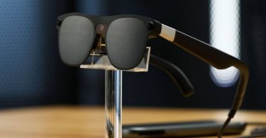 Lunettes de réalité mixte Xreal Project Aura de couleur noire exposées sur un support transparent. La monture, équipée de caméras et capteurs frontaux, est reliée par un câble à un boîtier externe posé sur la table, illustrant le fonctionnement filaire nécessaire pour alimenter les écrans OLED et l'écosystème Android XR.