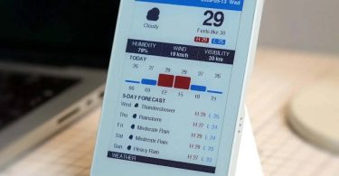 Boîtier M5Paper Color ESP32S3 affichant une interface météo colorée à Shenzhen sur son écran e-ink de 4 pouces, posé sur un support blanc à côté d'un ordinateur portable.
