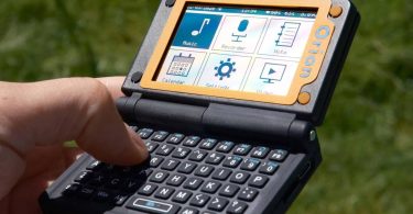Un utilisateur tient en main l'Orion PDA, un assistant numérique au format clapet, ouvert en extérieur. L'écran monochrome affiche une interface épurée avec des icônes (Calendar, Settings). Le clavier physique complet QWERTY est visible au premier plan, et le clapet supérieur intègre un panneau solaire photovoltaïque.