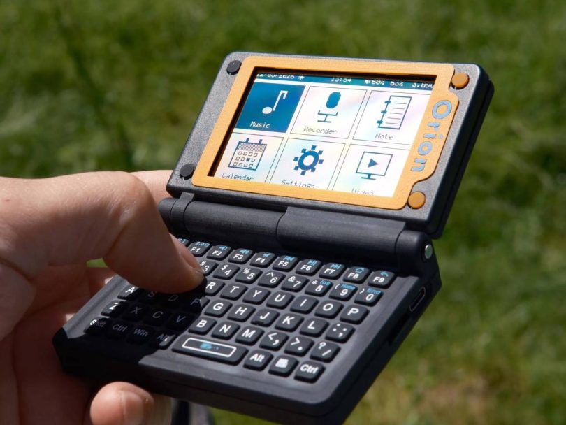 Un utilisateur tient en main l'Orion PDA, un assistant numérique au format clapet, ouvert en extérieur. L'écran monochrome affiche une interface épurée avec des icônes (Calendar, Settings). Le clavier physique complet QWERTY est visible au premier plan, et le clapet supérieur intègre un panneau solaire photovoltaïque.