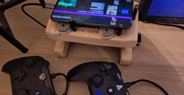 Vue rapprochée d'un smartphone Google Pixel 8, affecté par une ligne rose verticale sur son écran, posé sur un support en bois fait main. L'écran du téléphone affiche l'interface d'EmulationStation (ES-DE). Au premier plan, deux manettes de jeu filaires (style Xbox et rétro) sont branchées via un hub USB-C, prêtes pour une session de jeu.