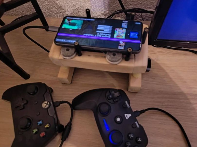 Vue rapprochée d'un smartphone Google Pixel 8, affecté par une ligne rose verticale sur son écran, posé sur un support en bois fait main. L'écran du téléphone affiche l'interface d'EmulationStation (ES-DE). Au premier plan, deux manettes de jeu filaires (style Xbox et rétro) sont branchées via un hub USB-C, prêtes pour une session de jeu.