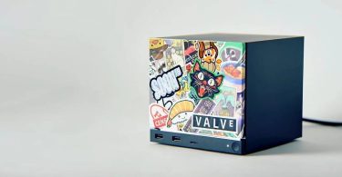 Mini PC Valve Steam Machine noir de forme cubique, posé de trois-quarts face, dont la façade avant est entièrement recouverte de stickers colorés et de graffitis de style pop-culture, incluant un logo officiel "VALVE" en bas à droite et des ports USB en façade.